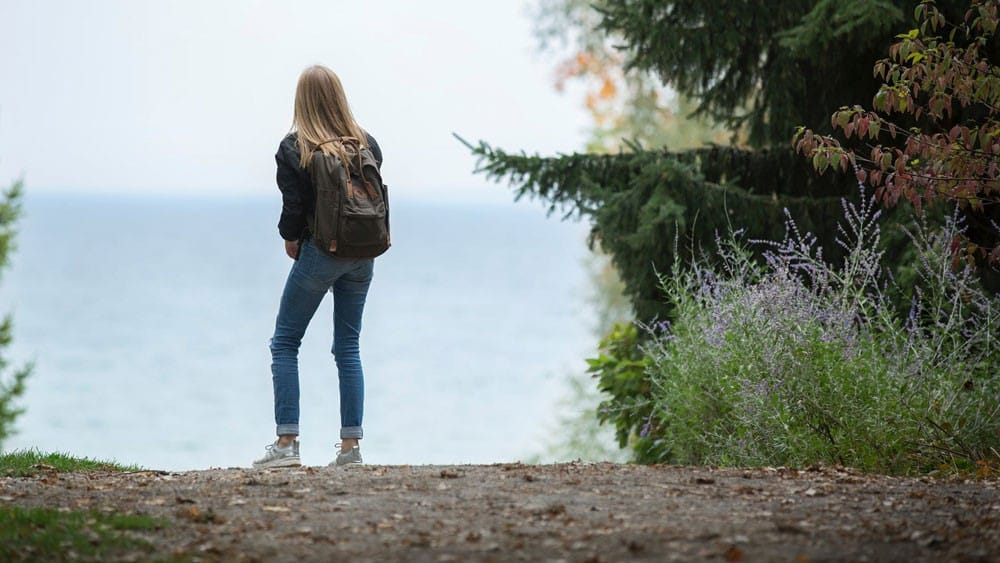 lugares increíbles en Canadá Chica rubia de espaldas con vaqueros y chaqueta negra mirando al mar al lado de la vegetación